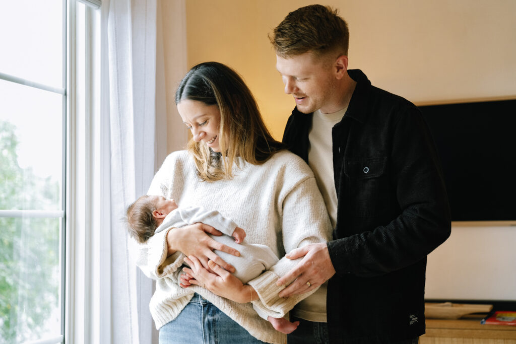 Mother and father smiling at their newborn by the window
