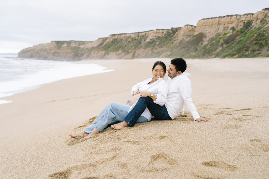 Bay Area maternity session Half Moon Bay — couple sitting on sandy shore with ocean and cliffs