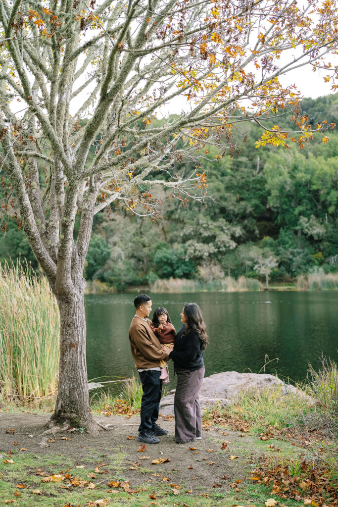 Family photography at Boronda Lake Bay Area — family of three standing by lake with autumn tree and green hills