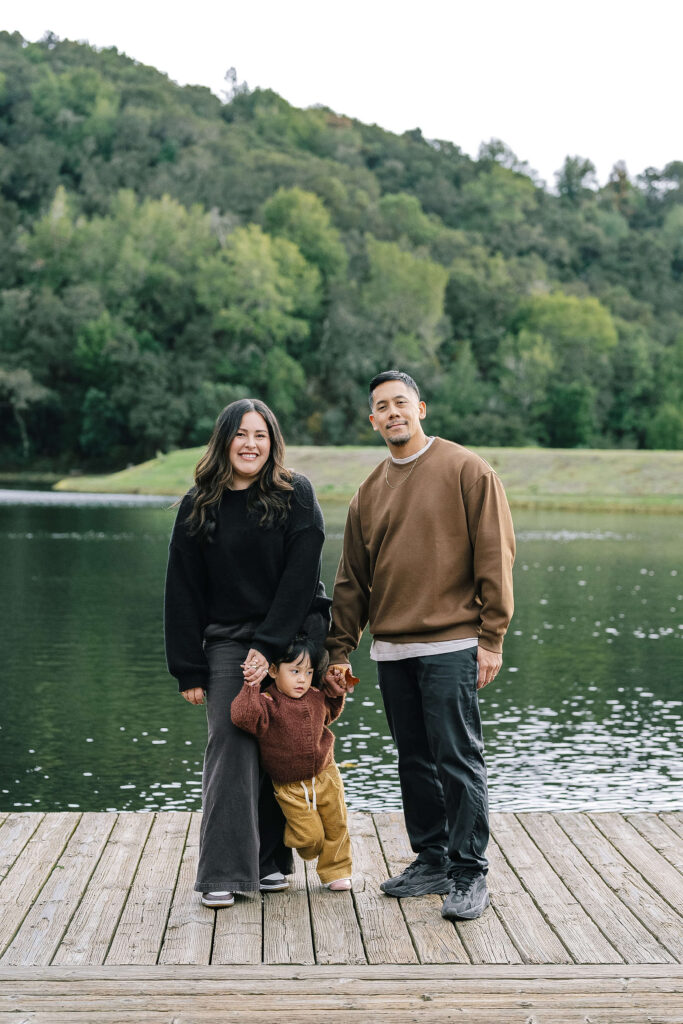 Lifestyle family session Boronda Lake South Bay — family with toddler smiling on wooden dock with lake and hills