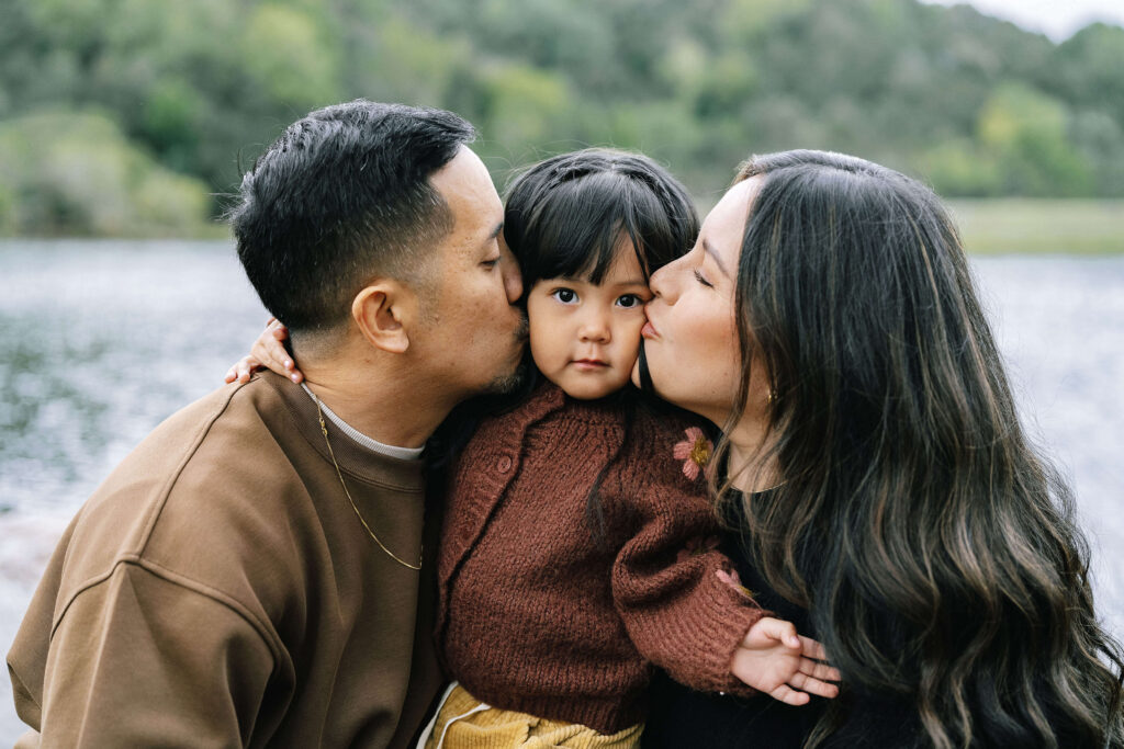 Candid family portrait Boronda Lake Bay Area — parents kissing toddler on cheeks with lake and green hills in background