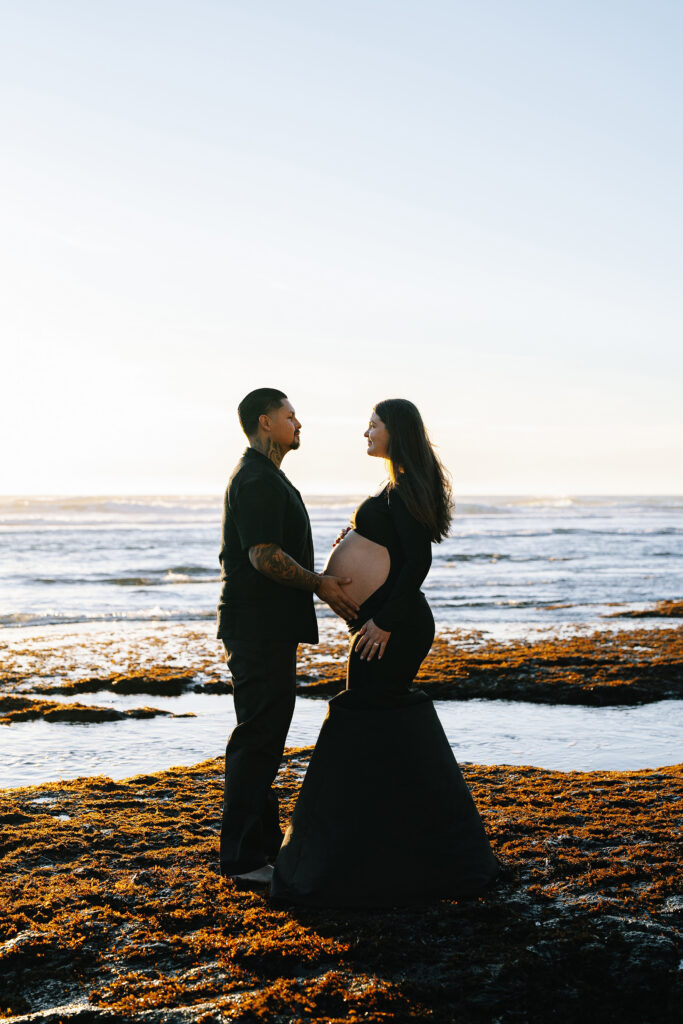 Maternity session at Moss Beach — couple facing each other on rocky shore at sunset Bay Area