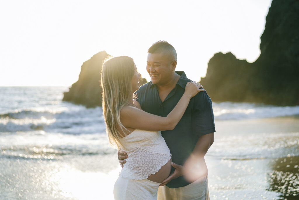 Intimate maternity portrait Rodeo Beach Marin Headlands — couple embracing with golden sunset backlight and ocean waves