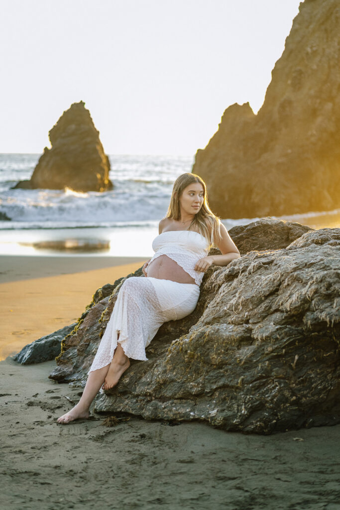 Maternity photography Rodeo Beach California — pregnant woman in white dress sitting on rock at sunset with coastal cliffs