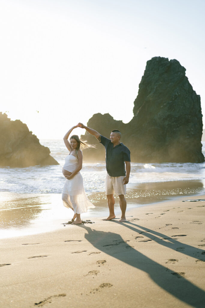 Maternity session at Rodeo Beach Marin Headlands — couple dancing on sandy shore at golden hour with dramatic rock formations